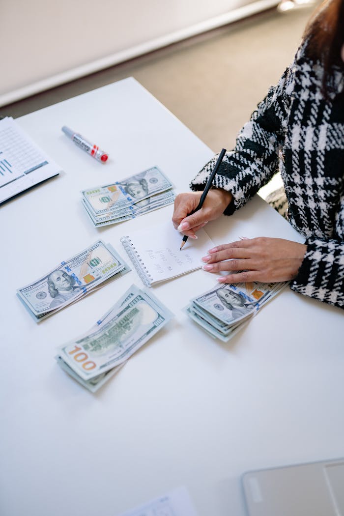 About A woman calculating finances with piles of cash and a notepad on a white table.